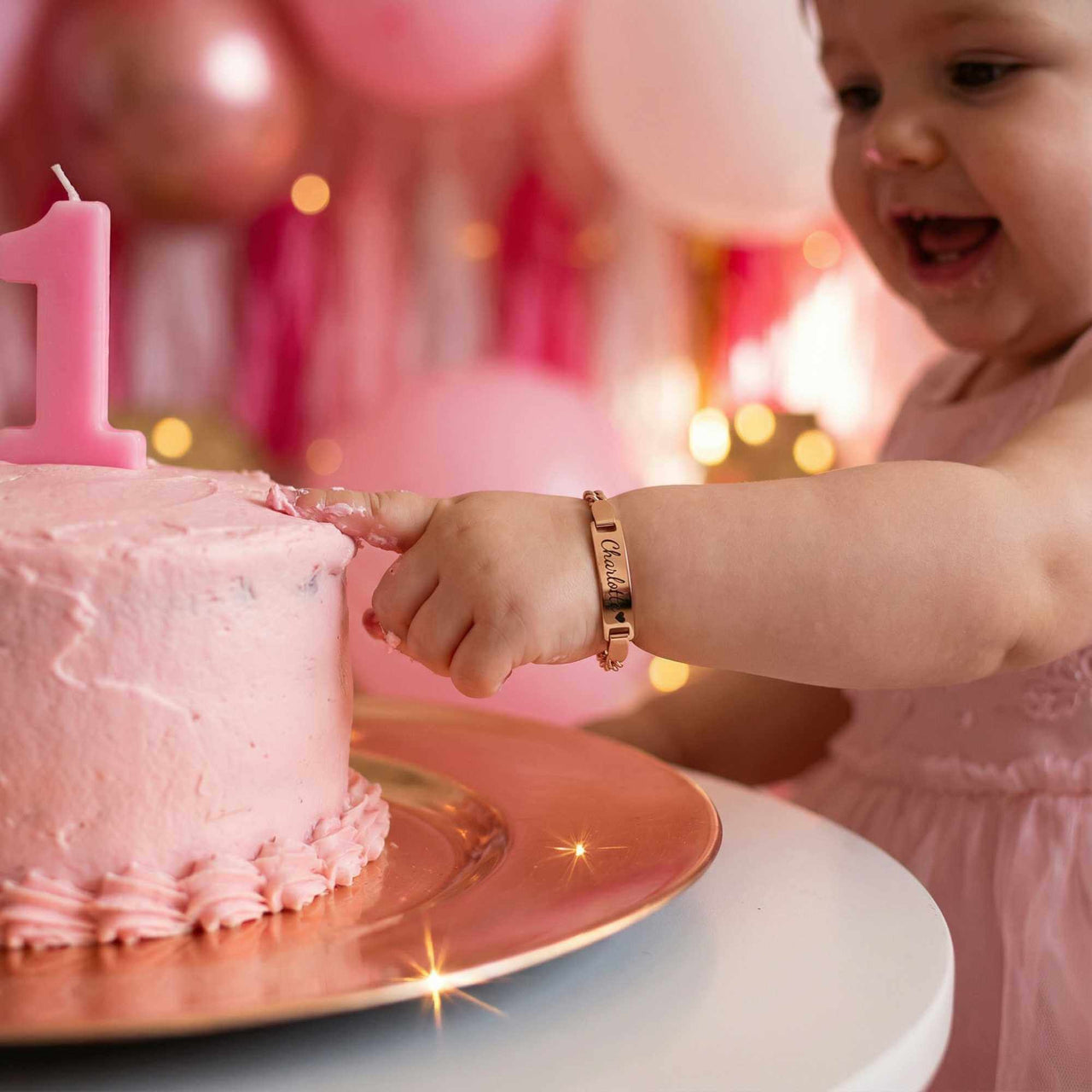 Infant Bracelet worn by a joyful baby reaching for a pink birthday cake, celebrating their first birthday with Brother & Sisters.