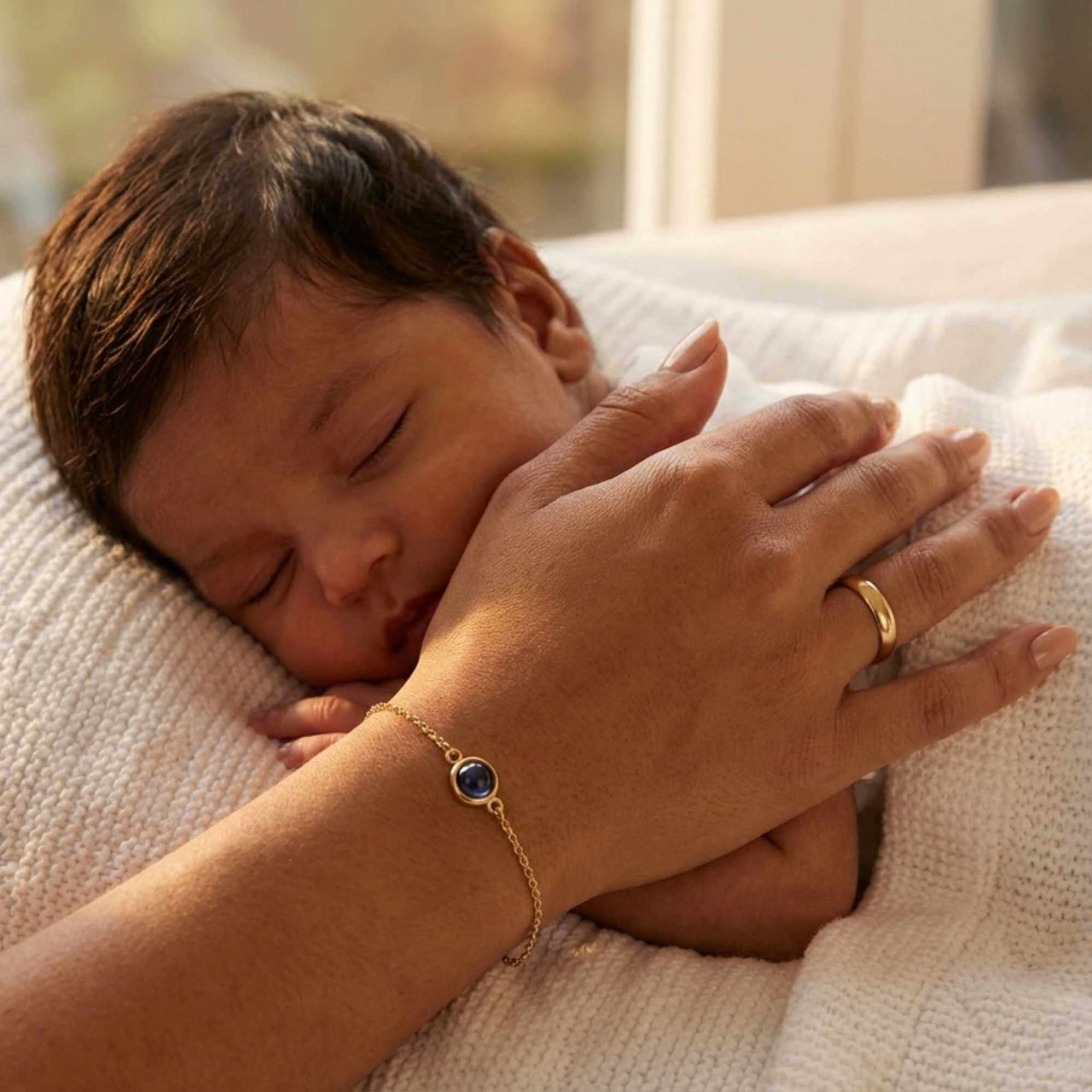 A close-up of a person's hand wearing a Photo Projection Bracelet by Brother & Sisters, with a sleeping baby nearby.