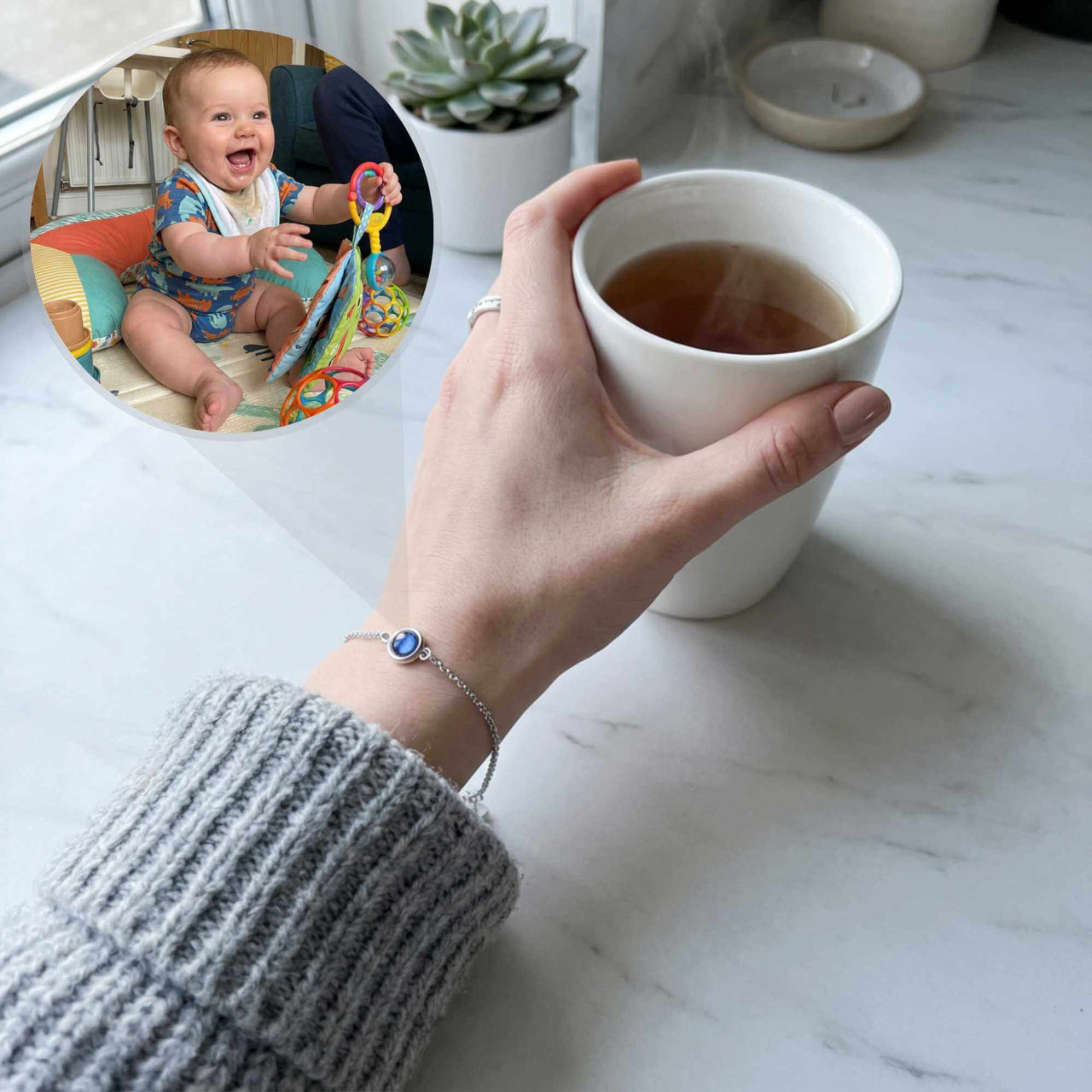 Hand holding tea cup while wearing a Photo Projection Bracelet by Brother & Sisters, with a smiling baby in background.