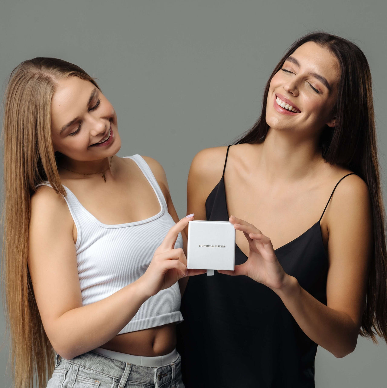Two smiling women holding a gift box from Brother & Sisters, showcasing the Heart Name Necklace.