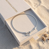 Thumbnail for Layla silver anklet inside B&S signature white slide-drawer gift box on beach sand at golden hour with sea shell — flat herringbone chain closed oval, fermoir and B&S tag visible, luxury silver anklet packaging