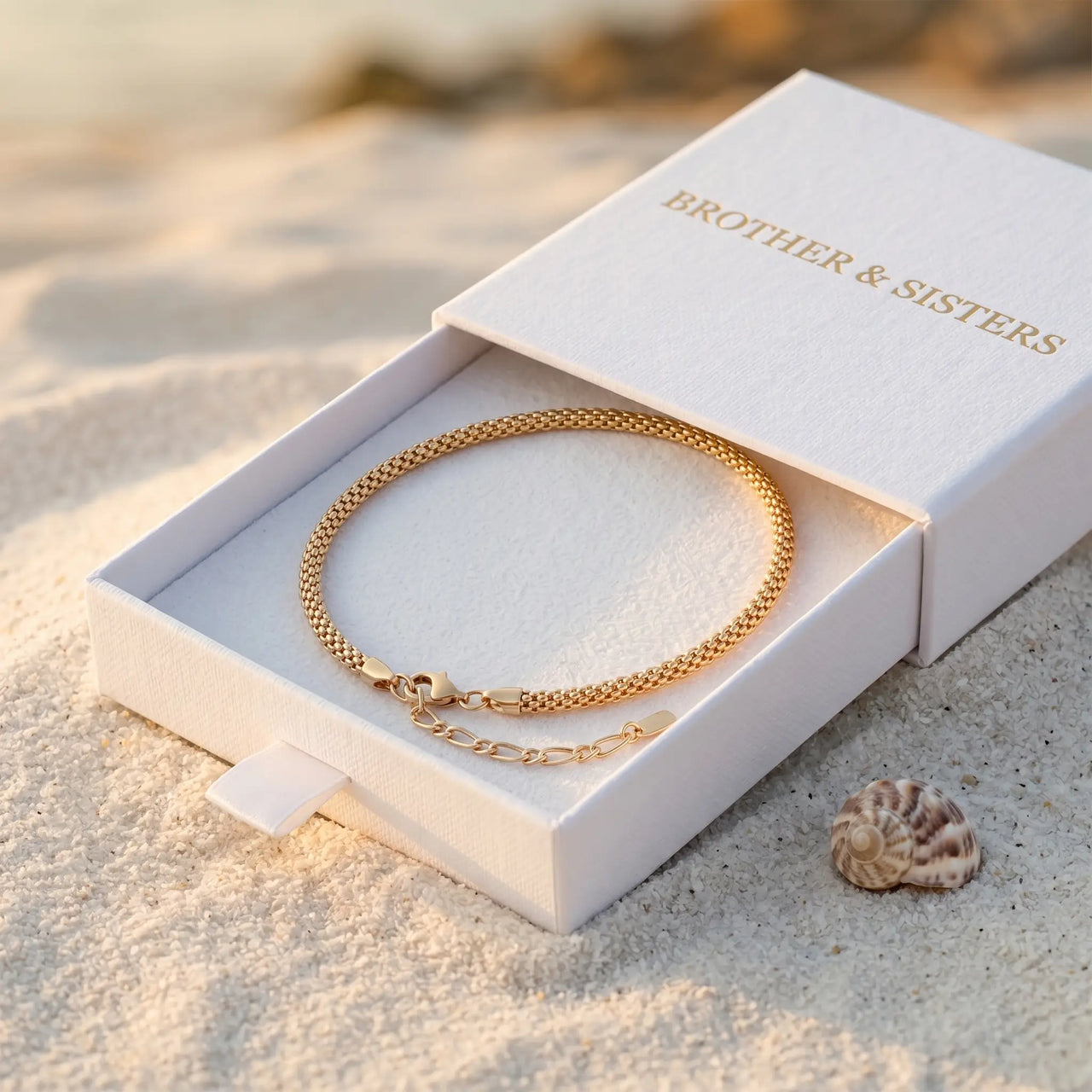 Maëlle gold tube-mesh anklet inside B&S signature white slide-drawer gift box on beach sand at golden hour with sea shell — luxury anklet packaging