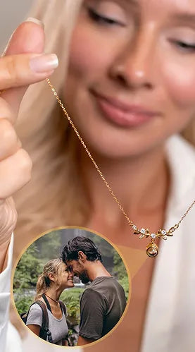 Blurred close-up of a blonde woman gently holding a fine gold necklace featuring a detailed vine-and-leaf pendant accented with clear cubic zirconia stones. The pendant includes a spherical charm that projects a personalized photo of a couple embracing outdoors, highlighting the necklace's memory feature.
