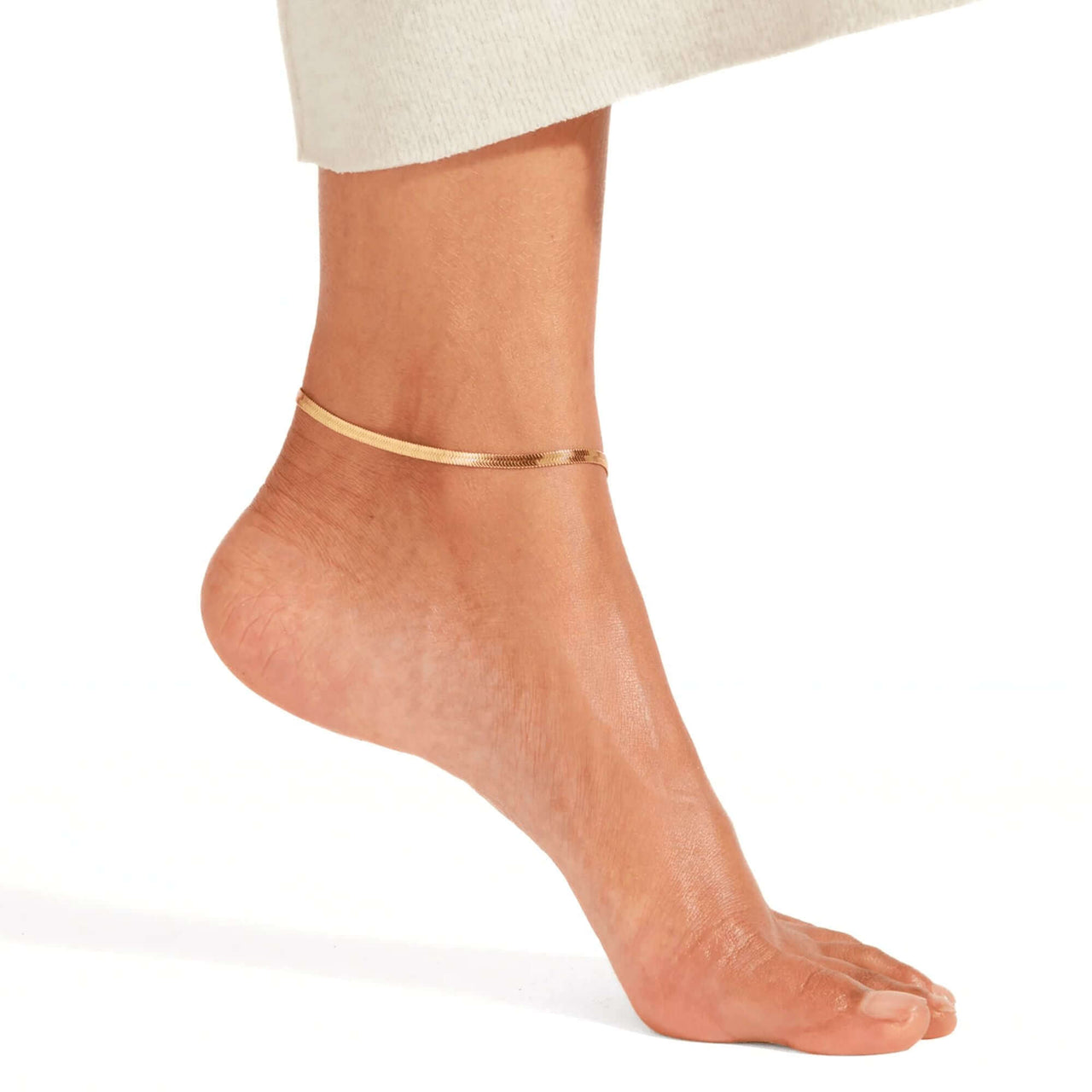 Layla gold herringbone snake chain anklet worn on tanned ankle with cream knit skirt — 18K gold-plated flat snake chain for everyday wear
