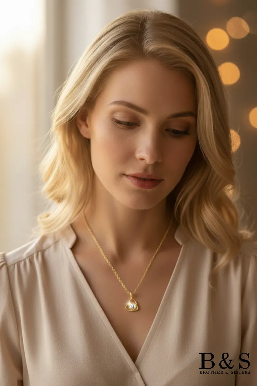 Professional studio shot of an elegant blonde model displaying a delicate gold-plated necklace with an open heart pendant and a central luminous pearl accent, set against a warm, soft-focus background.
